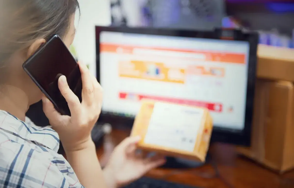 Osoba odwrócona tyłem rozmawia przez telefon trzymając paczkę przy monitorze ze sklepem online — Subiekt GT — obsługa zamówień telefonicznych i mailowych