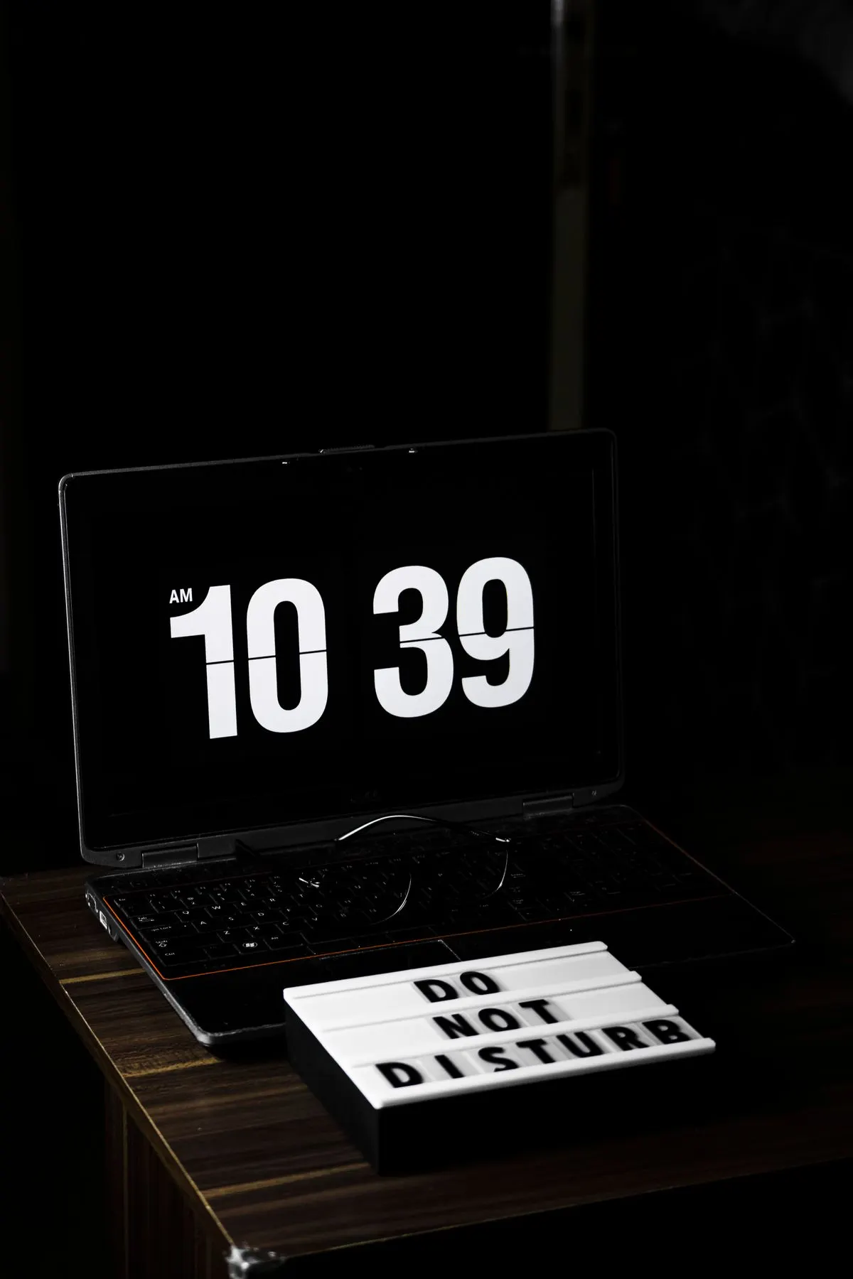 Laptop w ciemnym pomieszczeniu wyświetlający godzinę, obok tabliczka Do Not Disturb — Zdalna pomoc techniczna poza standardowymi godzinami pracy