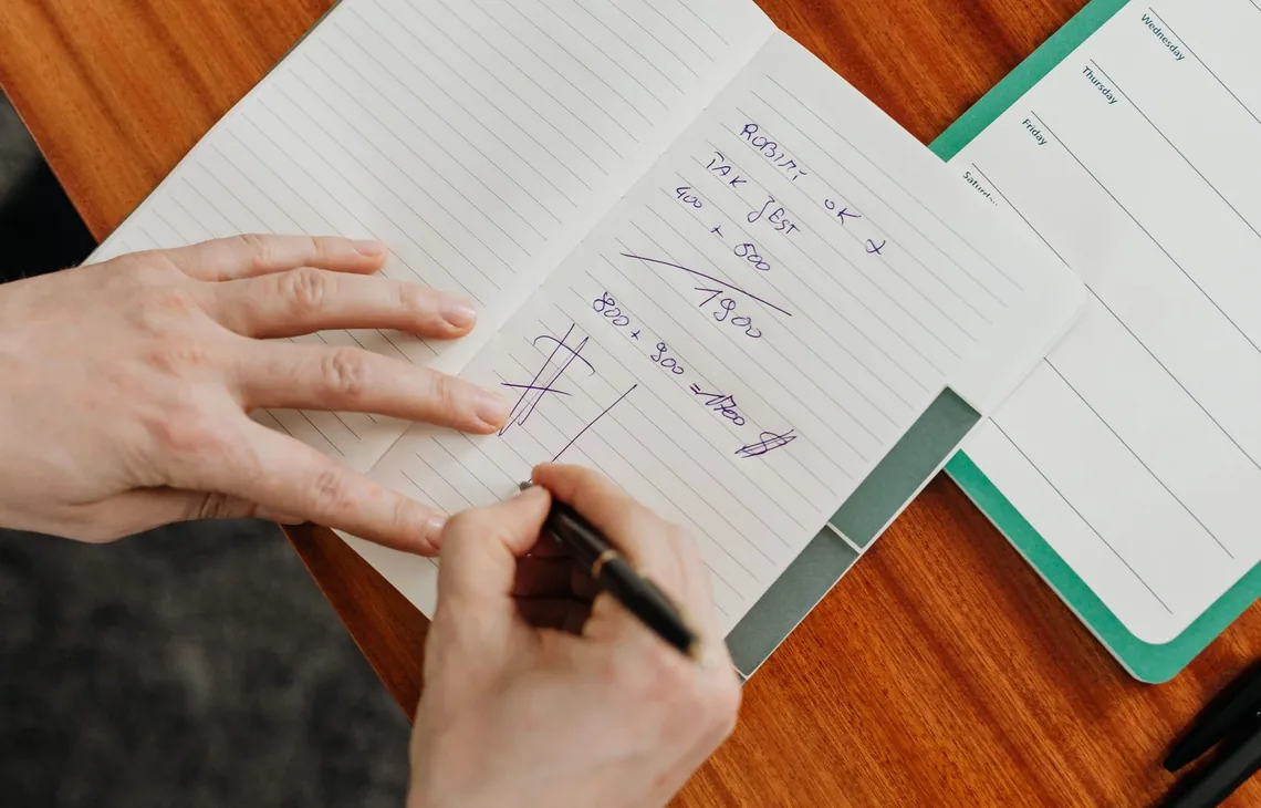 A person writing calculations in a notebook on a wooden desk.