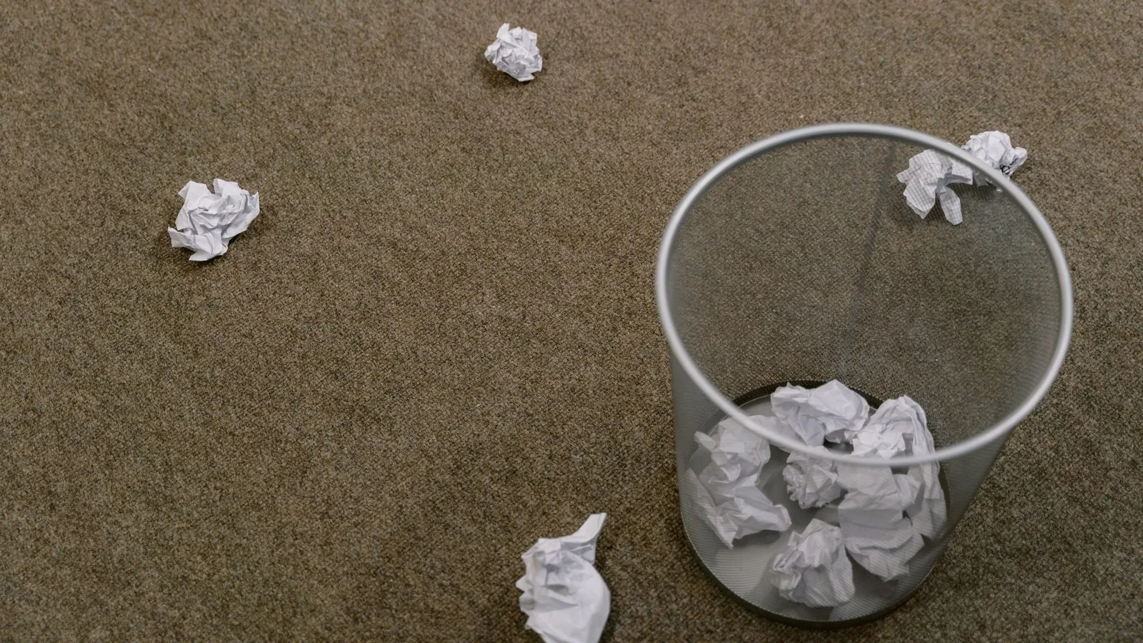 Crumpled paper scattered around a trash bin on a carpeted floor, indoors scene.