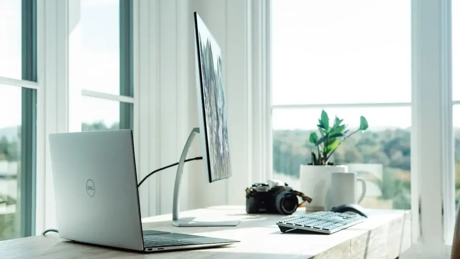 Srebrny laptop Dell przy biurku obok monitora, kamery i rośliny doniczkowej
