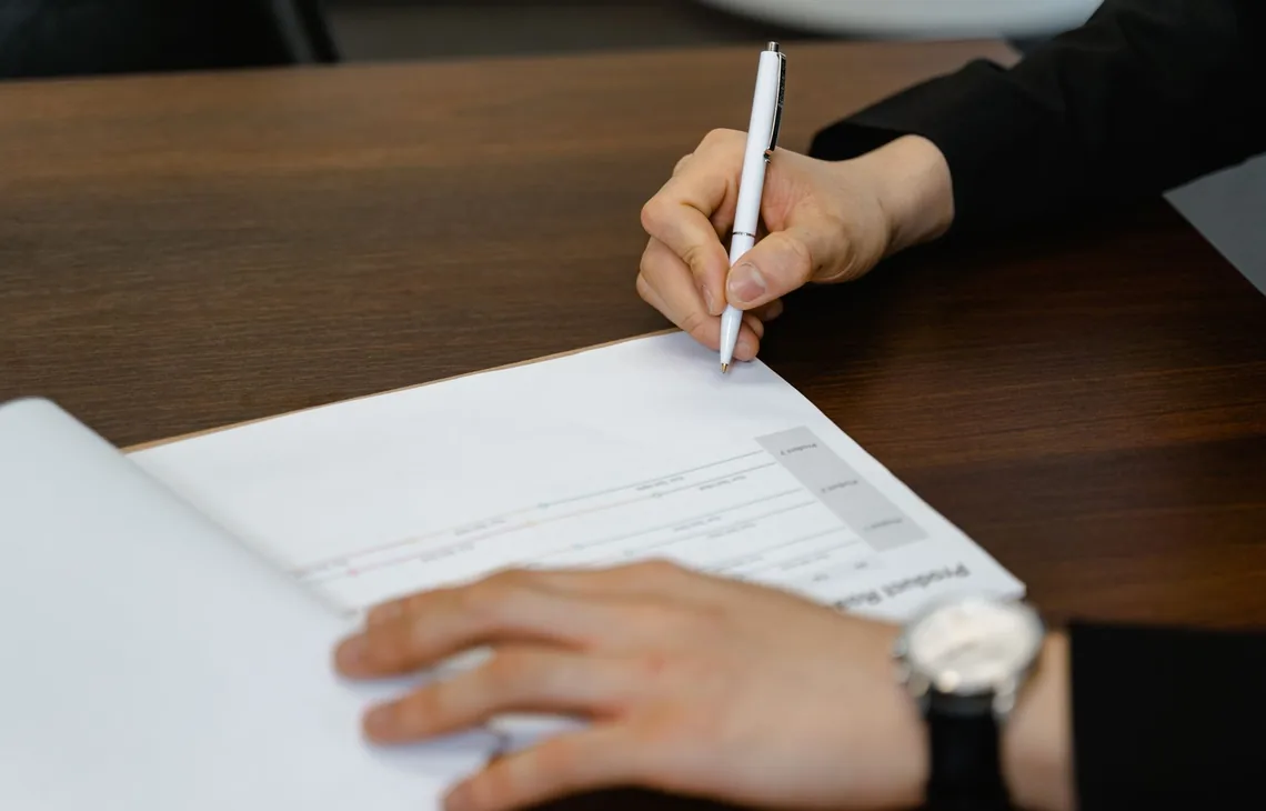 Businessperson writing on a document at a wooden desk, with focus on hands and pen.