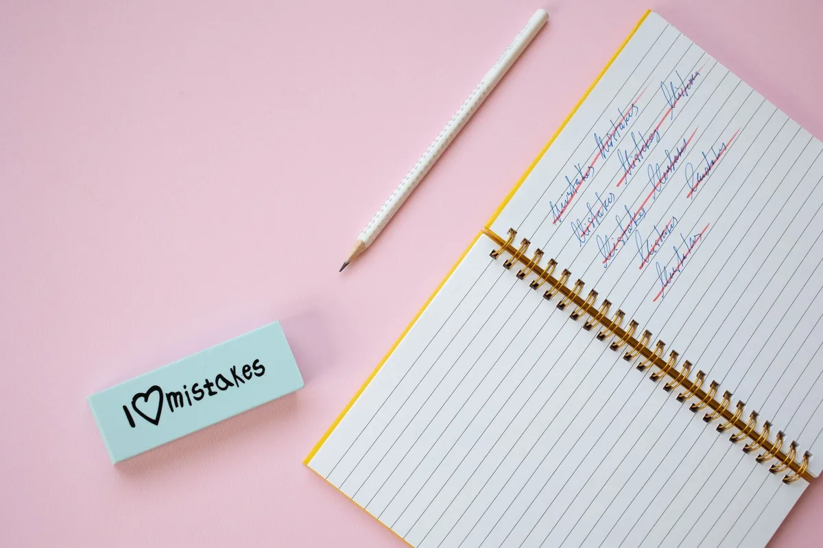 Flat lay of a spiral notebook and eraser on a pastel pink background with crossed out words.