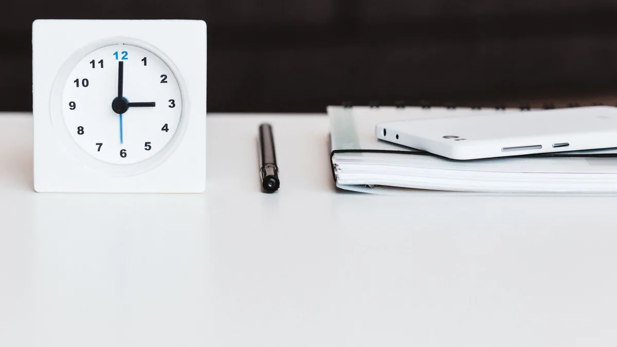 A minimalist office desk setup with a clock, pen, smartphone, and notebook, conveying time management and organization.