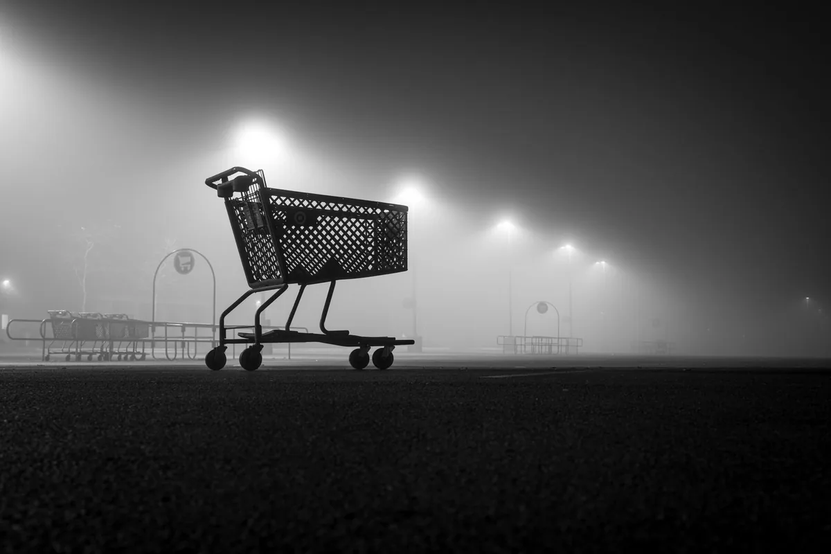 Black and white image of an empty shopping cart in foggy night parking lot.