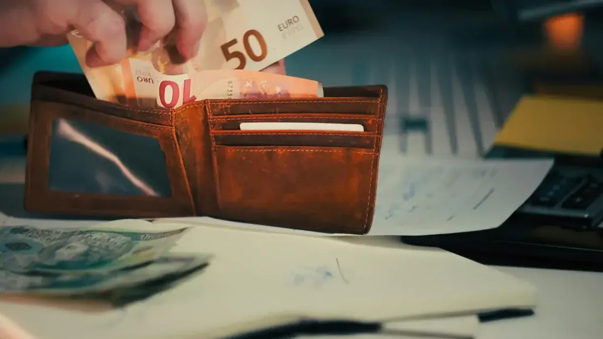 Dłoń wkładająca banknoty euro do portfela obok dokumentów i laptopa