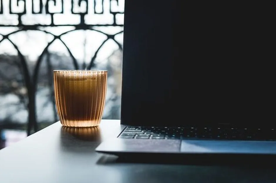 Otwarty laptop z kubkiem na stole przy oknie, wnętrze mieszkania o zmierzchu