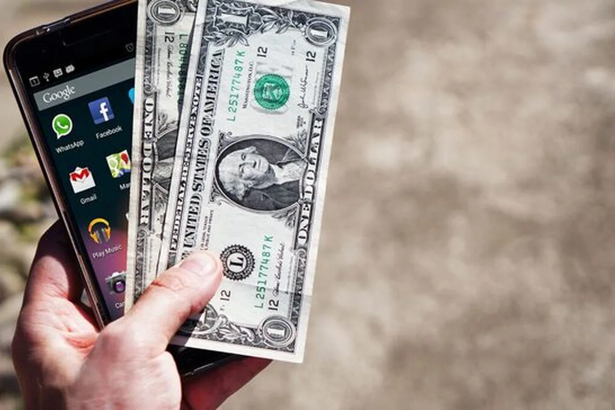 dłoń trzymająca smartfon i banknoty dolarowe