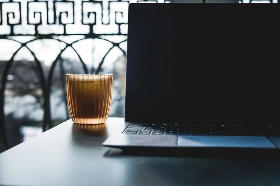 Otwarty laptop z kubkiem na stole przy oknie, wnętrze mieszkania o zmierzchu